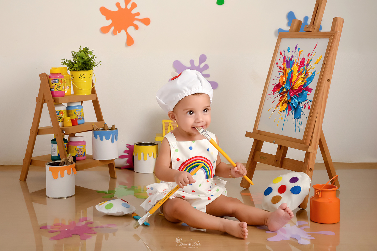 acompanhamento infantil - cenário de aquarela - dia das crianças - mês das crianças - cenários com latas de tintas - segurando dois pincèis - usando roupinha de pintura - e gorrinho na cabeça - marcas de tintas coloridas no chão e na parede 