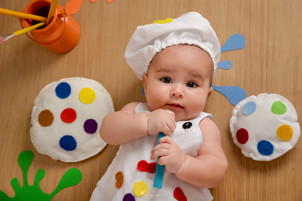 acompanhamento infantil - cenário de aquarela - dia das crianças - mês das crianças - cenários com latas de tintas - bebe deitado usando roupinha de pintor e gorrinho na cabeça - segurando pincel de pintura - 