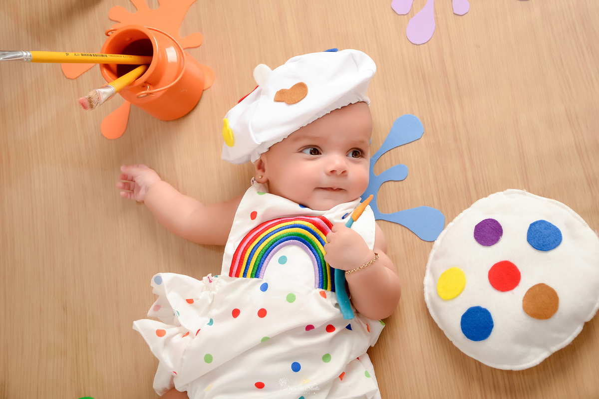 acompanhamento infantil - cenário de aquarela - dia das crianças - mês das crianças - cenários com latas de tintas - bebe deitada no cenário - usando roupinha de pintura - com gorrinho na cabeça - segurando pincel de pintura 