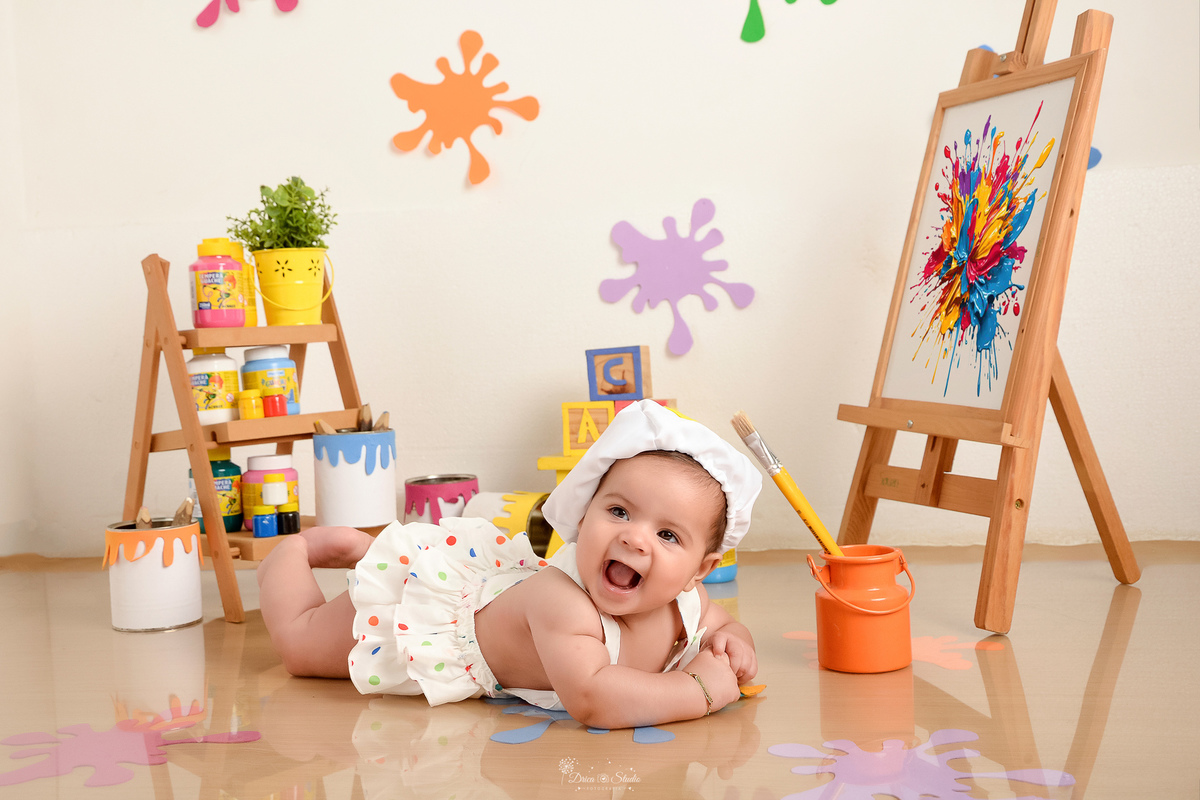 acompanhamento infantil - cenário de aquarela - dia das crianças - mês das crianças - cenários com latas de tintas e pinceis - marcas de tintas coloridas pelo chão e parede - bebe usando roupinha de pintora com gorrinho na cabeça - 