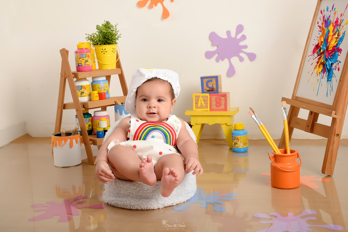 acompanhamento infantil - cenário de aquarela - dia das crianças - mês das crianças - cenários com latas de tintas - bebe sentanda em mini sofazinho - com os pezinhos levantados - usando roupinha de pintora e gorrinho na cabeça - 