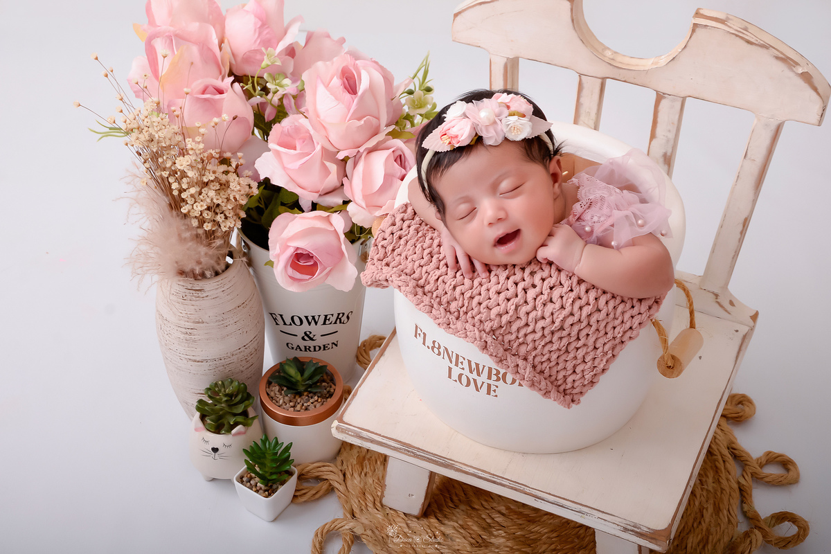 newborn - 11 dias de vida - primeiros dias de vida -  menina - baby - dentro de um mini baldinha - usando laço de flor rosa na cabeça - ao lados vasos de flores rosas - em cima de um baquinho branco de madeira - drica studio - xinguara - pará 