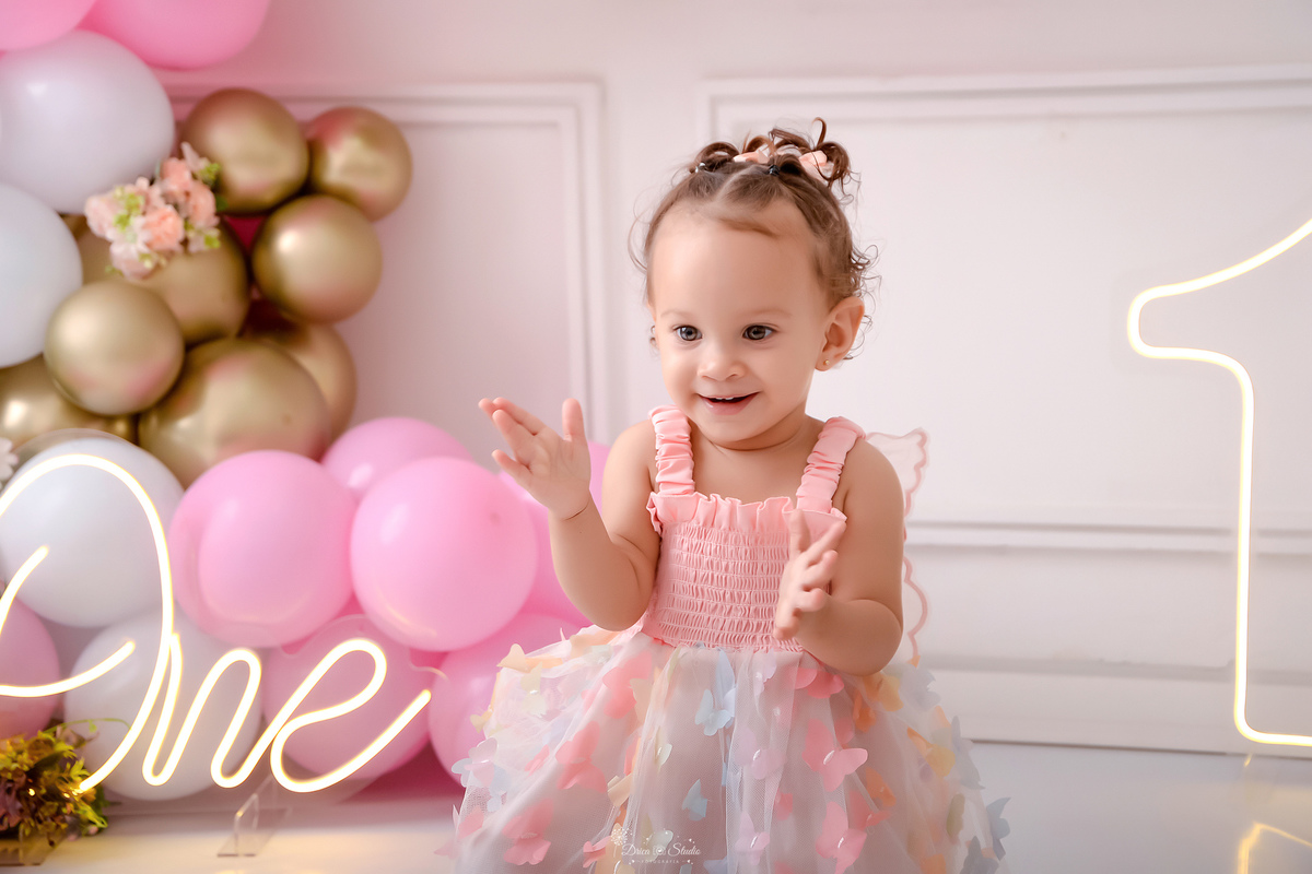 smash the cake - 1 ano - aniversário - cenário com balões nas cores branco, rosa e dourado - flores pelo cenário - 1 de led - one de led - bebê usando vestido rosa de borboletas - sentada em um baquinho branco - bebe batendo palmas - drica studio - pará