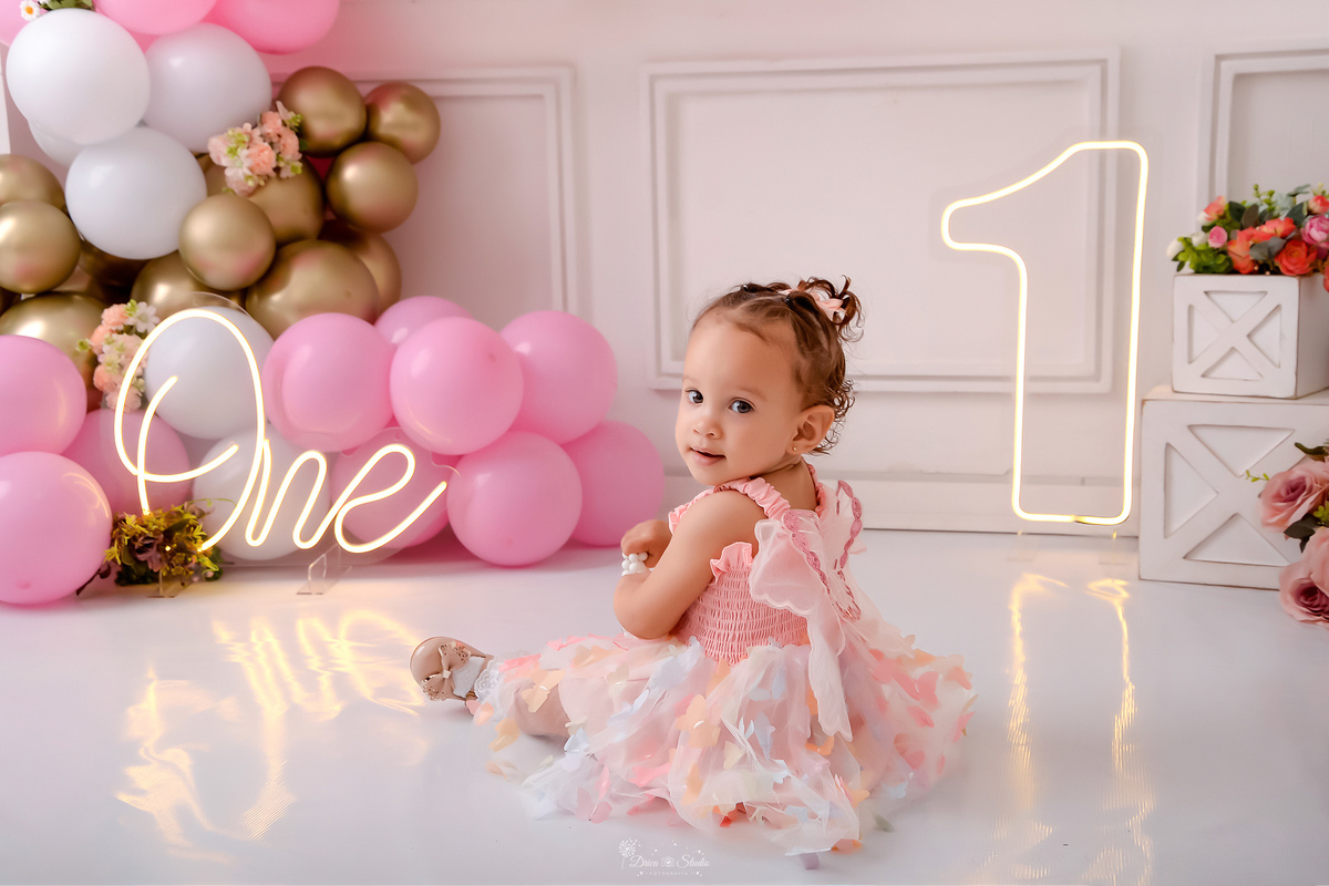 smash the cake - 1 ano - ensaio infantil - cenário com balões nas cores branco, rosa e dourado - flores pelo cenário - 1 de led - one de led - bebe sentada no cenário - usando vestido rosa de borboletas - drica studio - xinguara - pará - 
