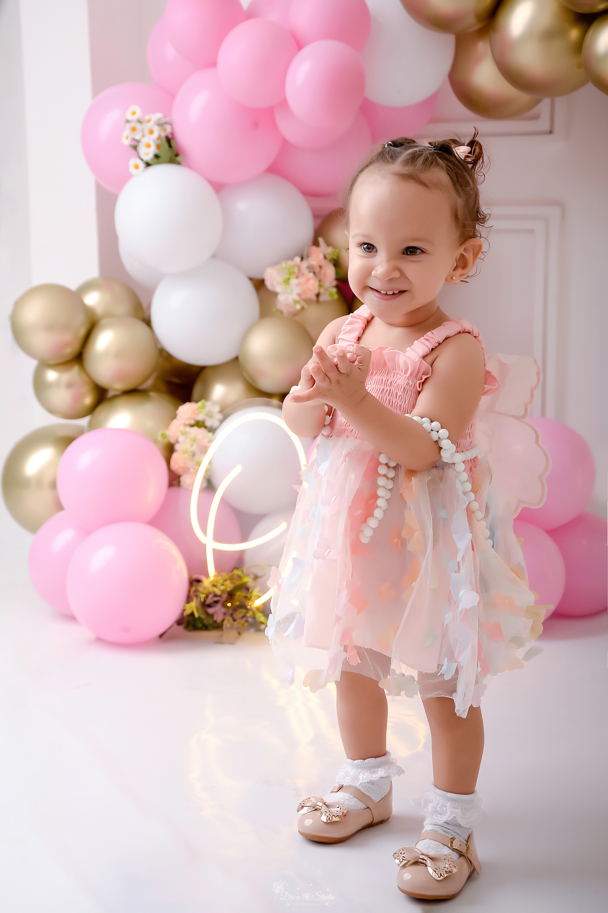 smash the cake - ensaio infantil - primeiro aninho - cenário com balões brancos, rosas e dourados - one de led - flores pelo cenário - bebe usando vestido rosa com borboletas - bebe batendo palmas - drica studio - xinguara - pará 