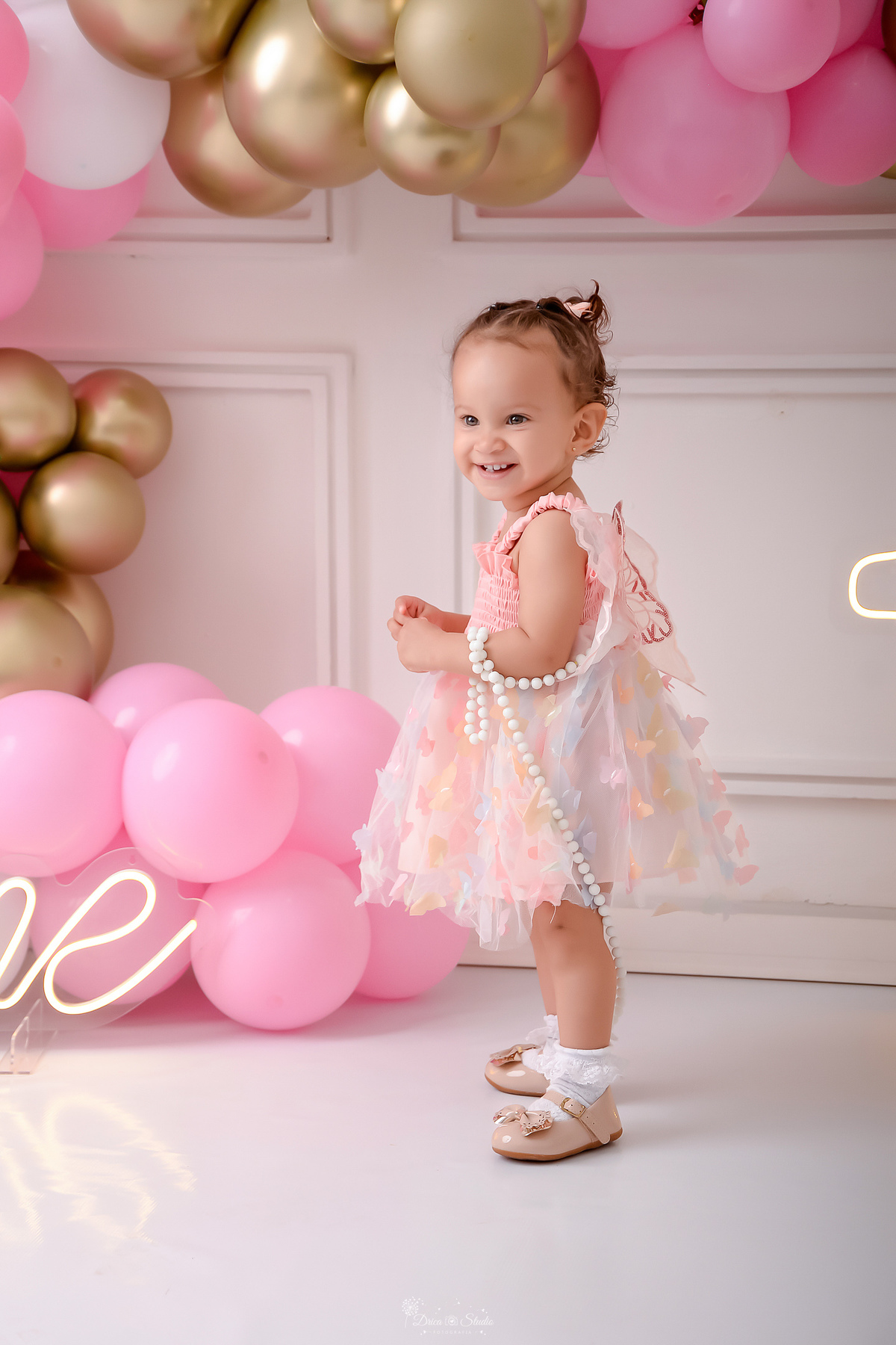 smash the cake - 1 aninho - ensaio infantil - 1 led - one de led - cenário com balões nas cores brancos, rosas e dourados - flores pelo cenário - bebe usando vestido rosa de borboletas - bebe em pé no cenário - drica studio - xinguara - pará - 