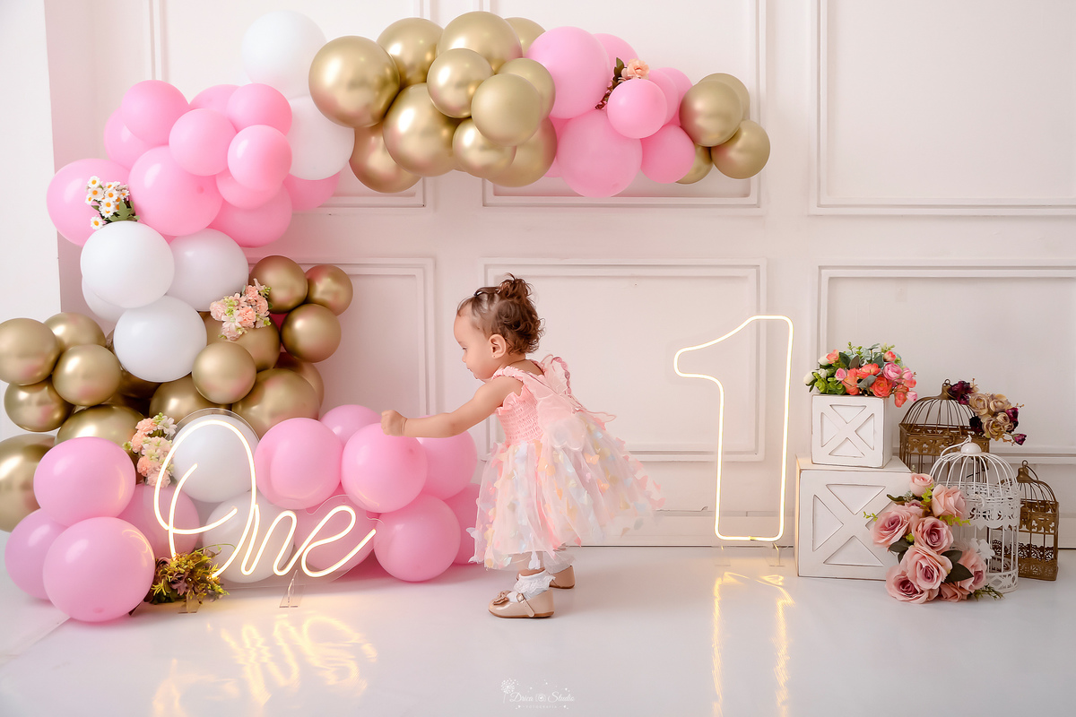 smash the cake - 1 aninho - ensaio infantil - primeiro aninho - cenário com balões nas cores - brancos - rosas e dourados - flores pelo cenário - 1 de led - one de led - bebe usando vestido rosa de borboletas - brincando com os balões - drica studio -