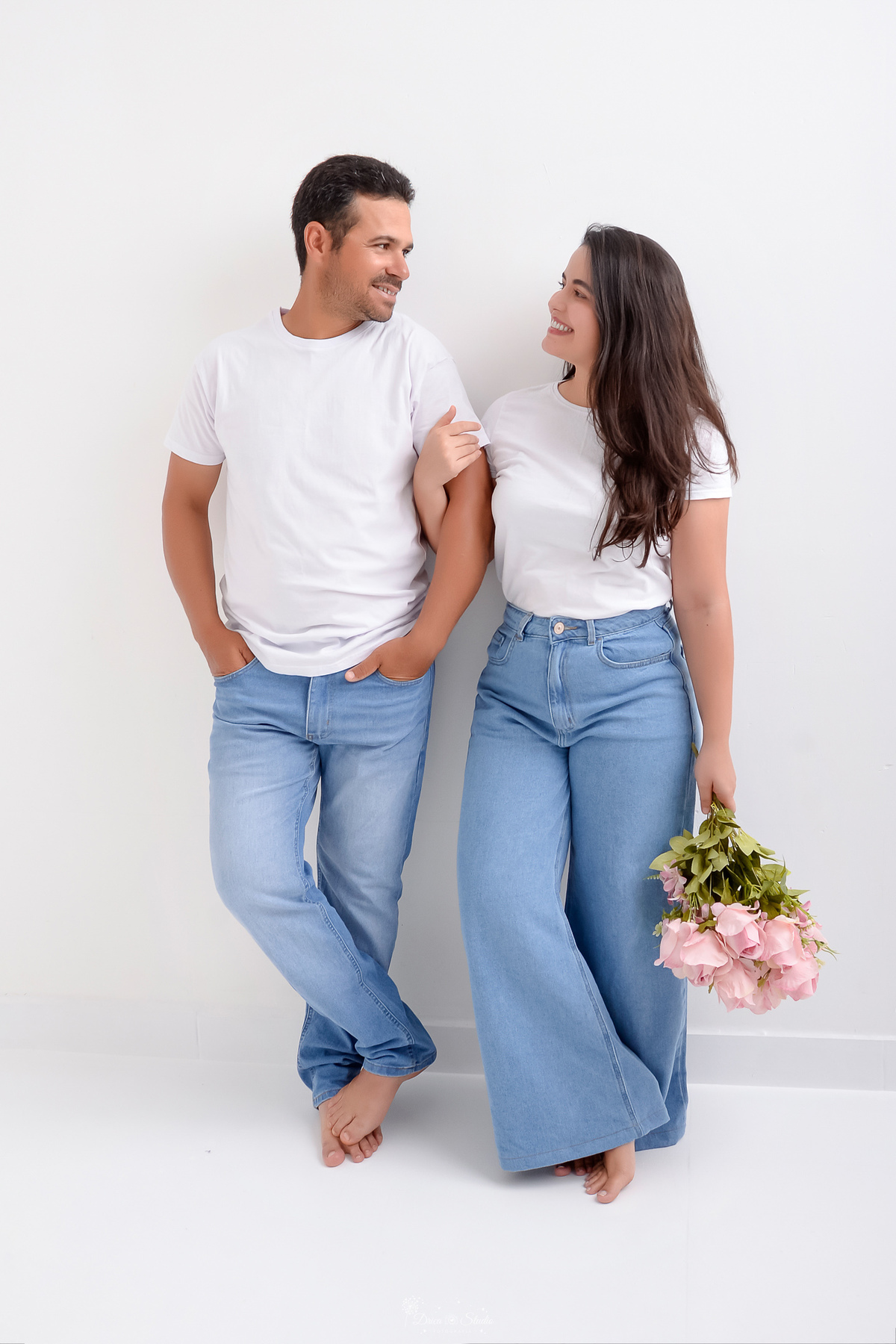 Ensaio casal - ensaio fotografico - fotos em estudio - drica studio - xinguara - pará - casal encostado em uma parede branca - usando camisas brancas e calça jeans clara - esposa segurando um buque de rosas claras - casal se olhando -