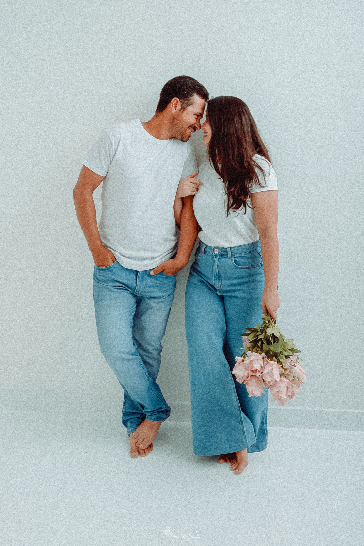 Ensaio casal - ensaio fotografico - fotos em estudio - drica studio - xinguara - pará - casal encostado em uma parede branca - usando camisas brancas e calça jeans - esposa segurando um buque de rosas claras - casal se beijando - 