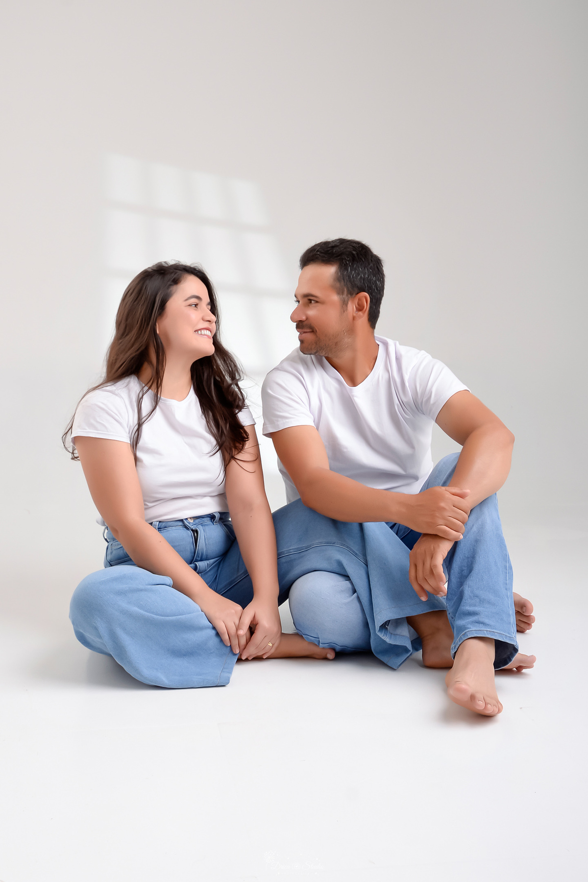 Ensaio casal - ensaio fotografico - fotos em estudio - drica studio - xinguara - pará - casal sentados no chão se olhando - usando camisas brancas e calça jeans - foto com fundo branco 