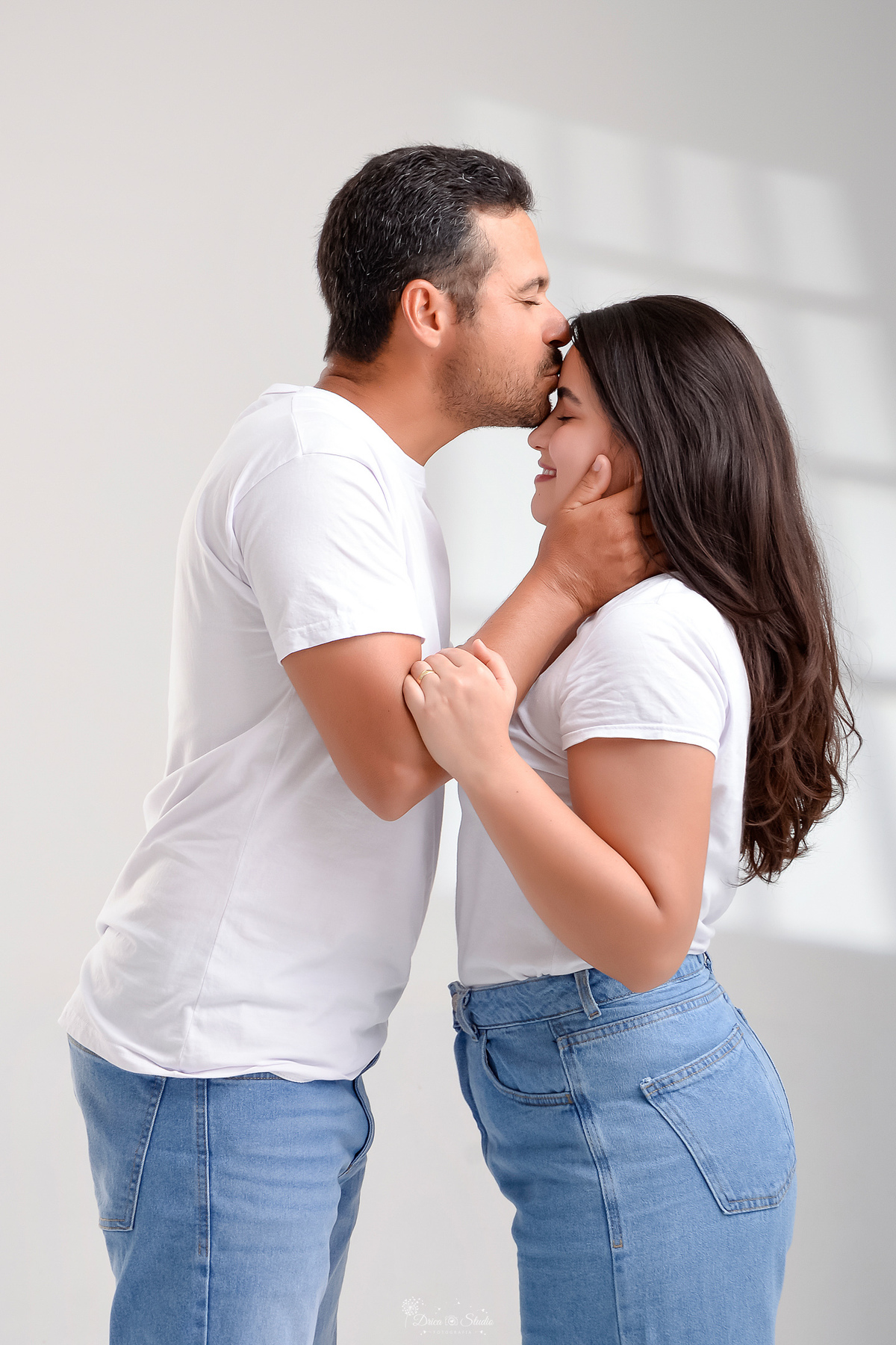 Ensaio casal - ensaio fotografico - fotos em estudio - drica studio - xinguara - pará - casal usando camisas brancas e calças jeans - esposo dando beijo na testa de sua esposa - esposa sorrindo olhando para seu marido - 