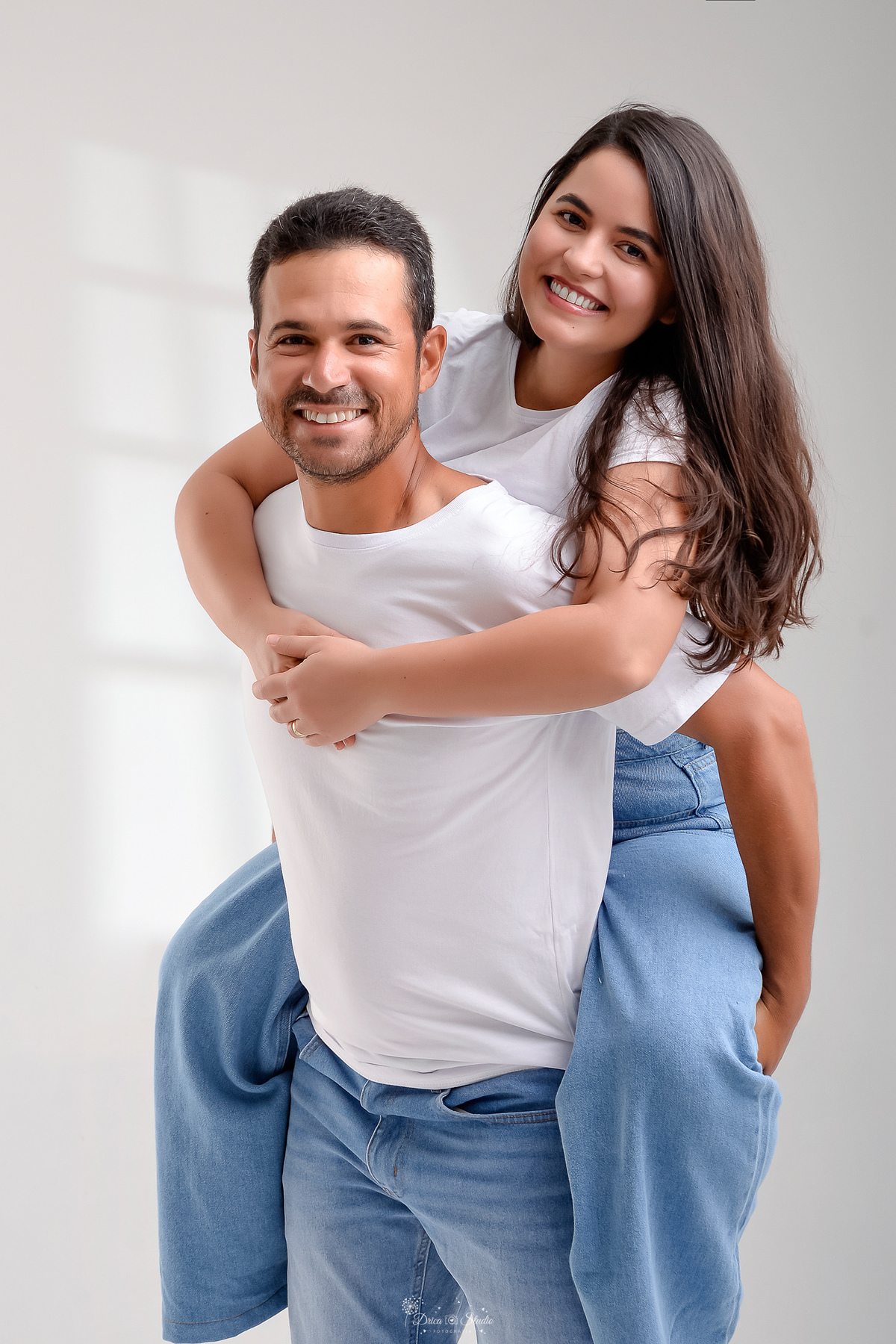 Ensaio casal - ensaio fotografico - fotos em estudio - drica studio - xinguara - pará - esposa sobre as costas do seu marido - casal usando camisas brancas e calça jeans - casal sorrindo para a foto - 