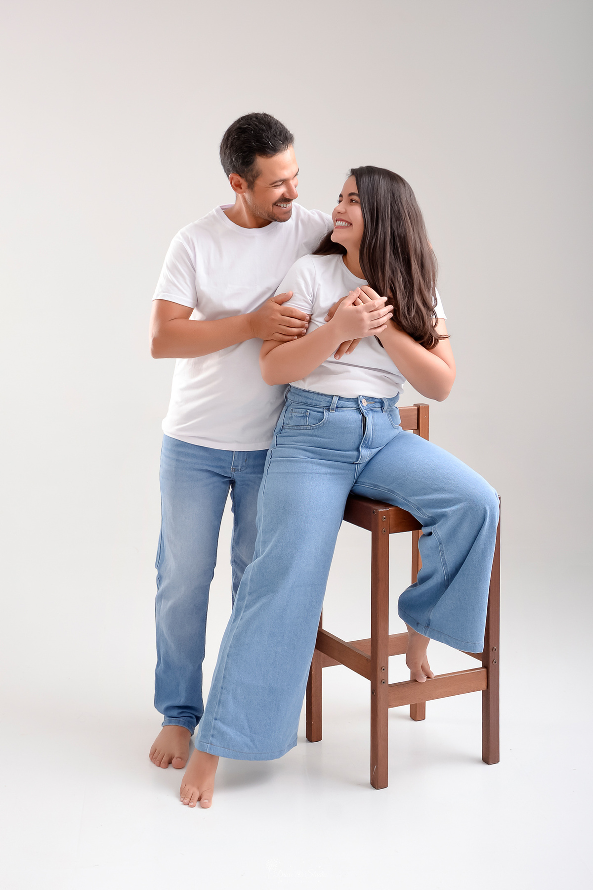 Ensaio casal - ensaio fotografico - fotos em estudio - drica studio - xinguara - pará - casal usando camisa branca e calça jeans clara - ela sentada em um banco de madeira e marido ao lado dela á abraçando 