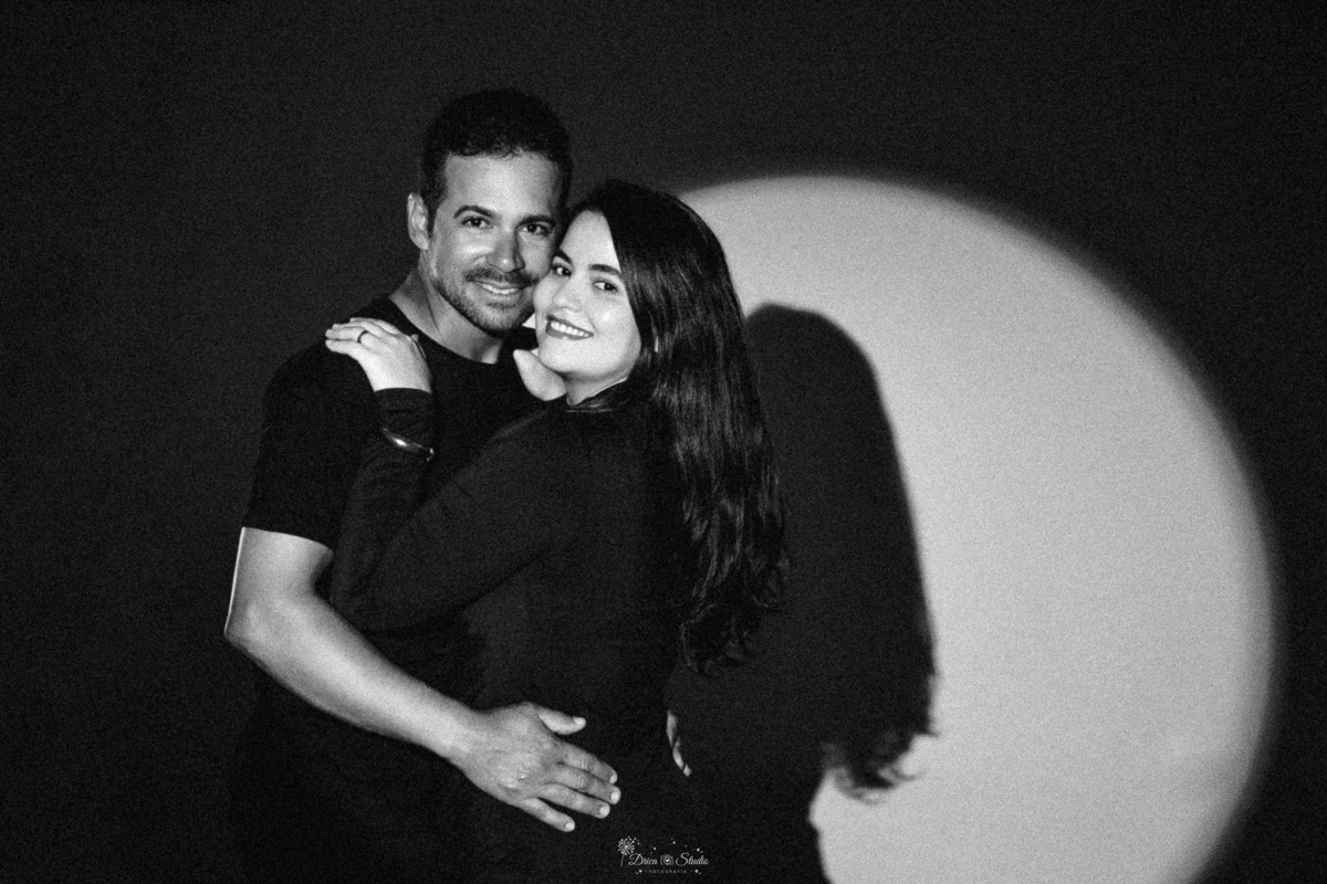Ensaio casal - ensaio fotografico - fotos em estudio - drica studio - xinguara - pará - casal abraçados - esposo usando camisa preta - esposa usando vestido de manga longa preto - luz ao fundo destacando as sobrars do casal - 