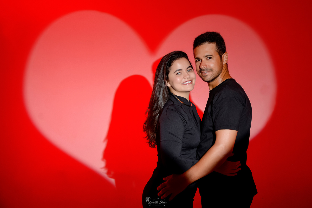 Ensaio casal - ensaio fotografico - fotos em estudio - drica studio - xinguara - pará - casal abraçados - esposo usando camisa preta e esposa usando vestido de manga longa na cor preta - ao fundo luz em formato de um coração - 