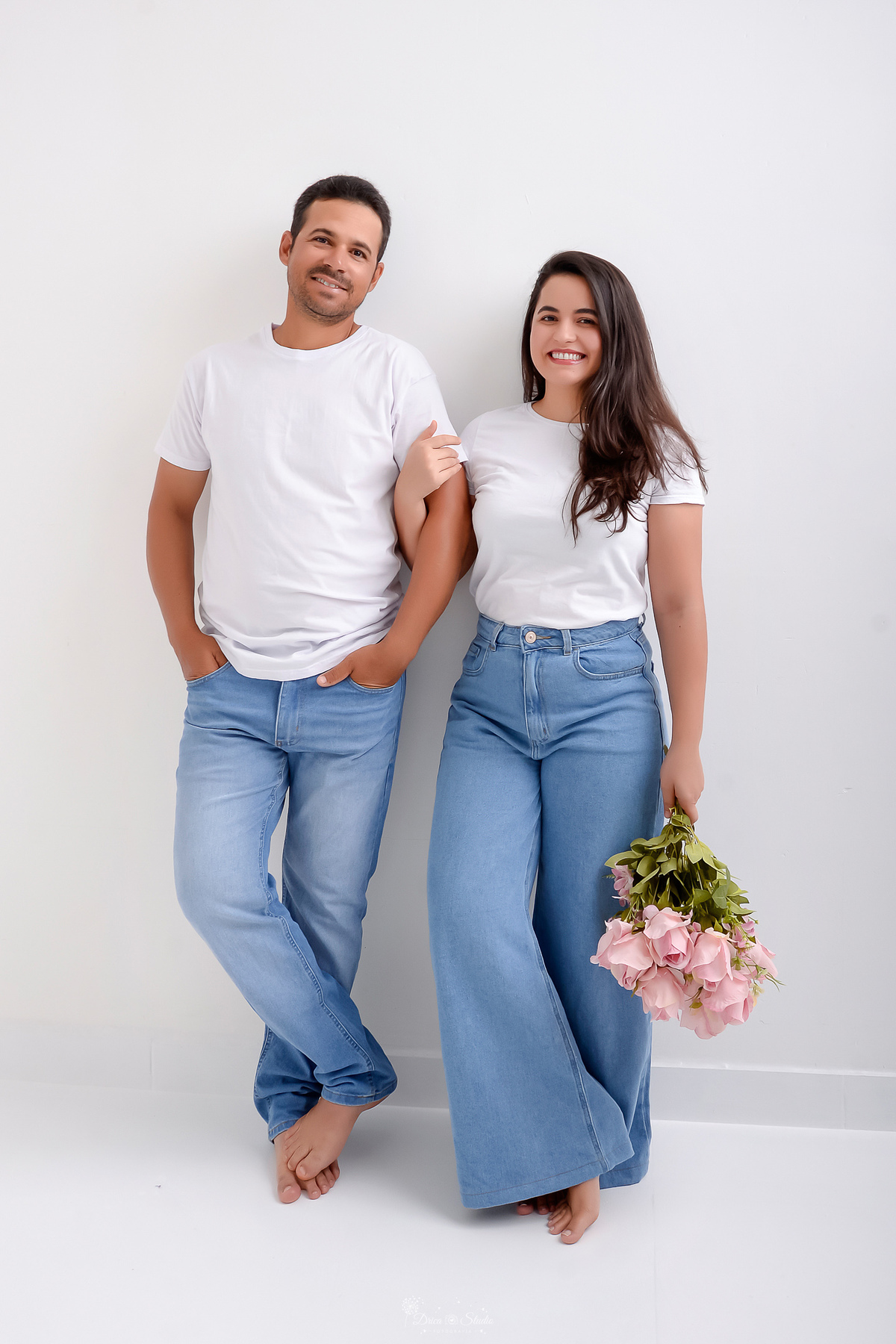 Ensaio casal - ensaio fotografico - fotos em estudio - drica studio - xinguara - pará - casal encostado na parede branca usando camisa branca e calça jeans - esposa segurando um buque de rosas claras   