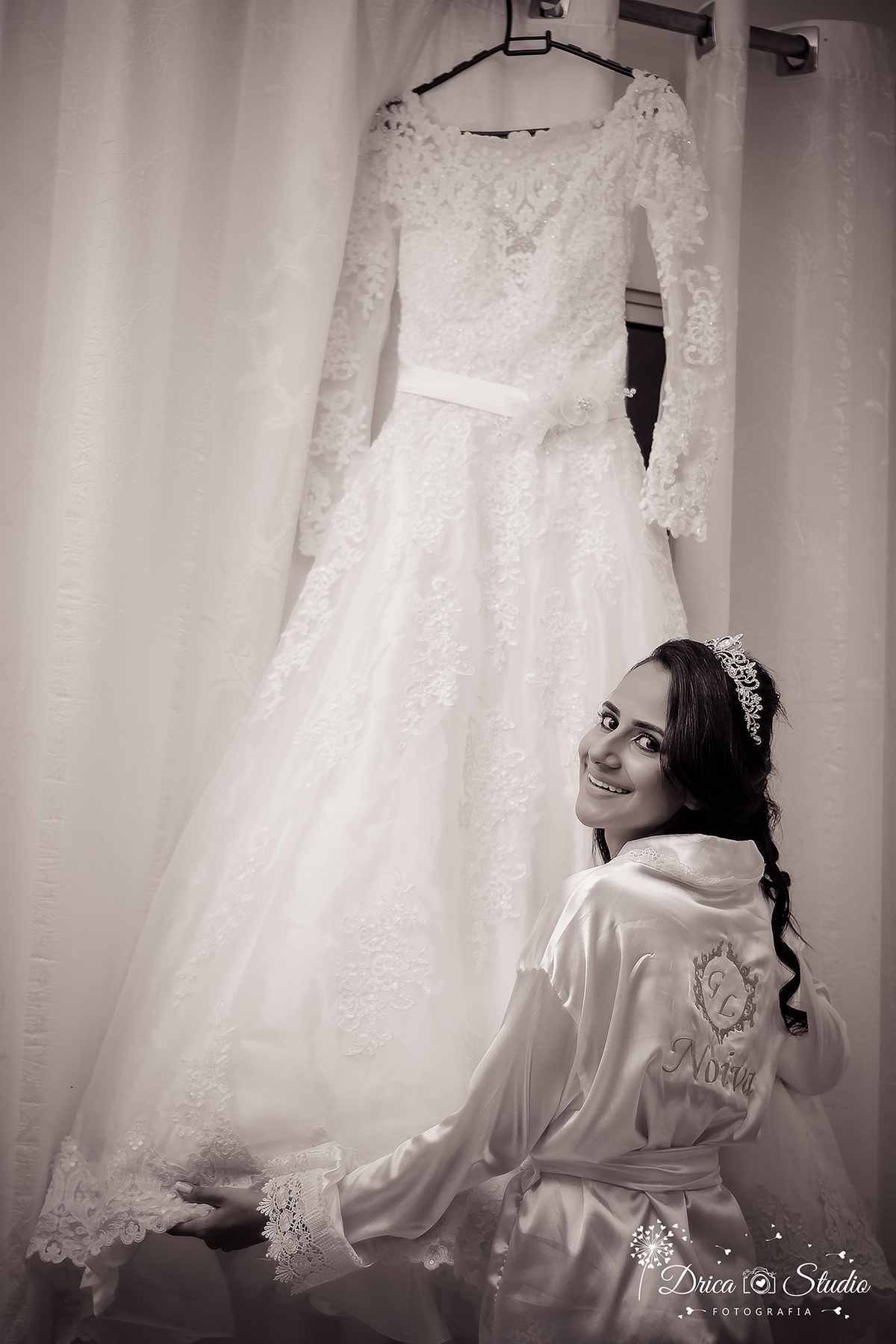 Making off foto preta e branca da Gabriela de roupão branco, com bordado de noiva nas costa ela olha sorrindo e abre seu vestido pendurado na janela fechada com cortinas brancas.