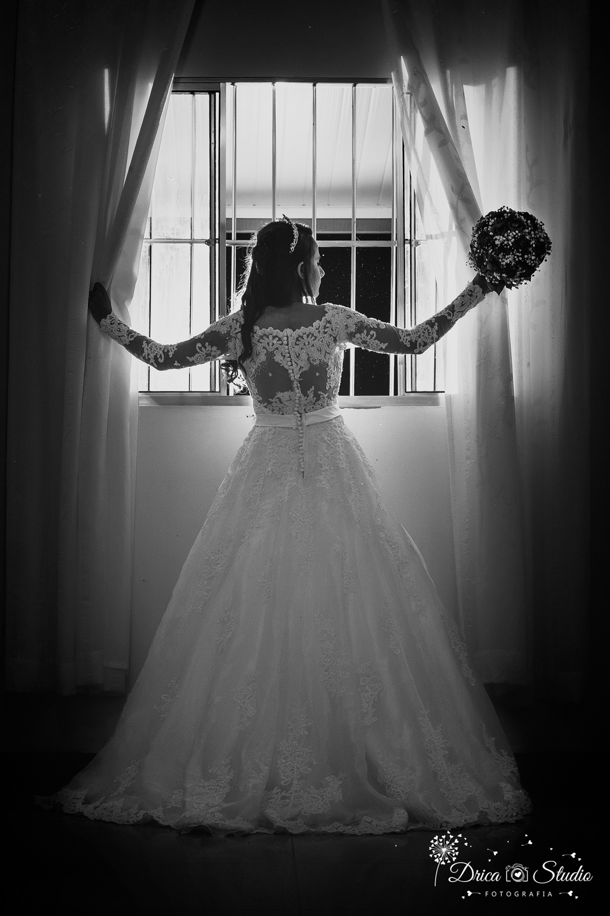 Making off foto preta e branca da noiva de costa olhando para o lado abrindo as cortina da janela contra luz com buquê em uma das suas mãos-Drica Studio-Fotografias-Fotógrafo de Casamento-Xinguara-Pará- Fotografa- Making of-Casamento-linda noiva-P&B.