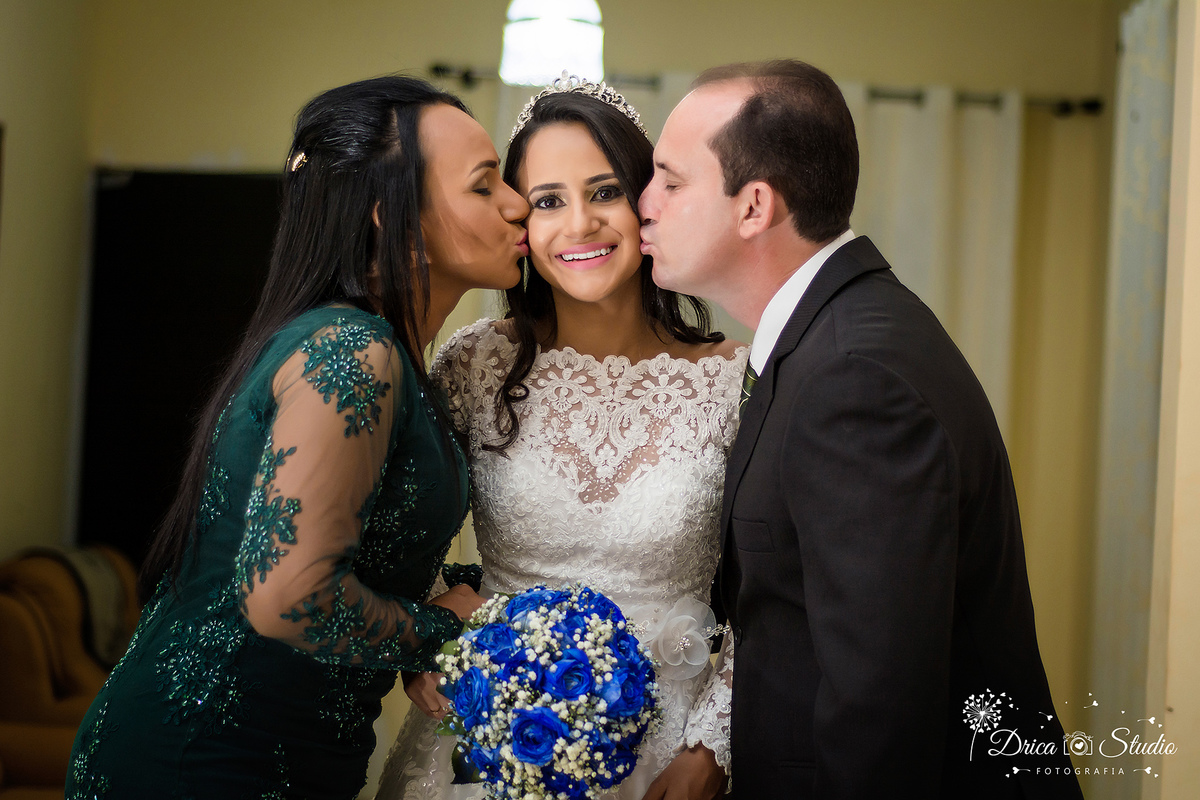 foto da nova sorrindo segurando seu buquê azul em uma sala amarelada sendo beijada pelo seus pais um de cada lado.Drica Studio-Fotografias-Fotógrafo de Casamento-Xinguara-Pará- Fotografa- Making of-Casamento-Sapucaia-Pá-Pais da Noiva-beijo-Amor-Verdadeiro