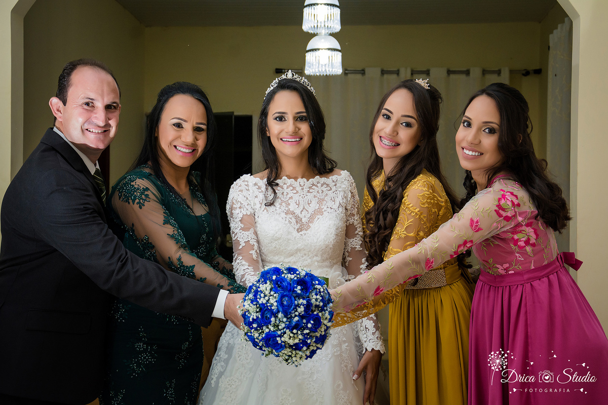 foto da noiva com seus pais de um lado e suas duas irmã do outro, lado bem trajados, sorridentes segurando seu buquê azul esticado para frente do seus corpo-Drica Studio-Fotografias-Fotógrafo de Casamento-Xinguara-Pará- Fotografa-Making of-Família- 