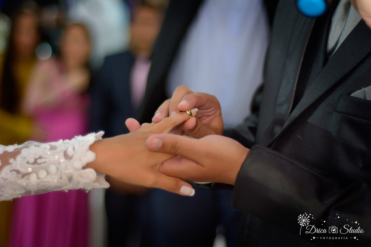 imagem só das mão dos  noivos em foco, ele colocando a aliança no dela dela com pessoas admirando ao fundo em desfoque.Drica Studio-Fotografias-Fotógrafo de Casamento-Xinguara-Pará- Fotografa- Casamento-Sapucaia-Pá-Aliança- Igreja- Cerimonia.