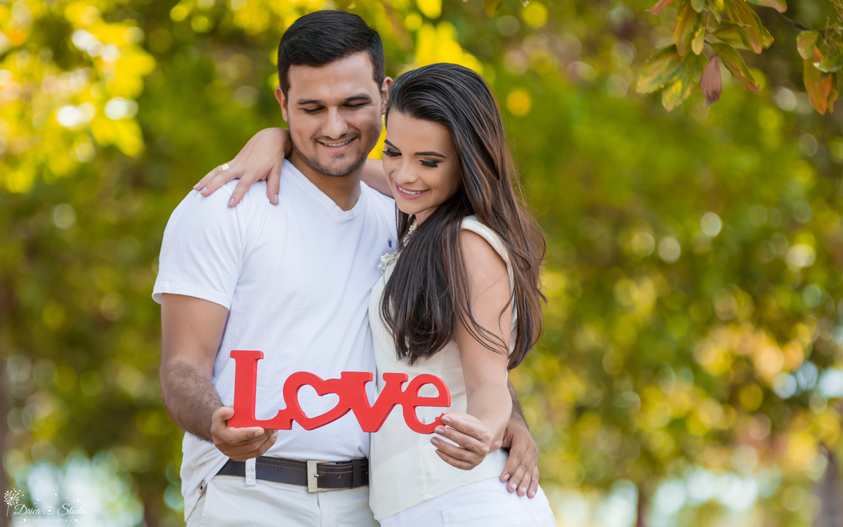 ensaio de casal abraçados segurando a palavra love na cor vermelha fundo com folhas em desfoque os noivos estão de branco em uma paisagem verde-Drica Studio- Fotografias- Fotografo- Casamento- Xinguara – Pará- Fotografa-Casal-Pre Wedding- Família-Externo.