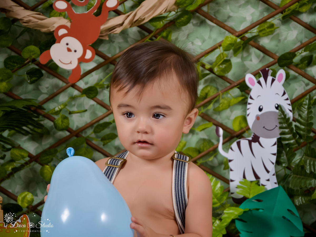 foto de uma criança em um senário safári segurando um balão azul, ao fundo uma tela verde com folhas verde e um desenho de zebra e do outro lado a de um macaco.Fotógrafa Infantil- Fotógrafo em Xinguara-Fotógrafo de Criança- Ensaio Infantil- Acompanhamento
