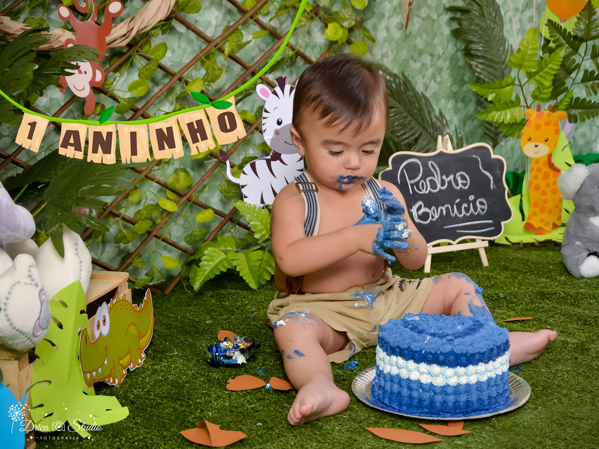 fotografia Smash da Criança com as mão toda melada de bolo na cor azul, onde ela a parenta abismada com a sensações. ao fundo a cor verde do tema safári chamam a atenção, as plaquinhas de animais quadrinho nome da criança. Drica Studio Fotografia-Xinguara