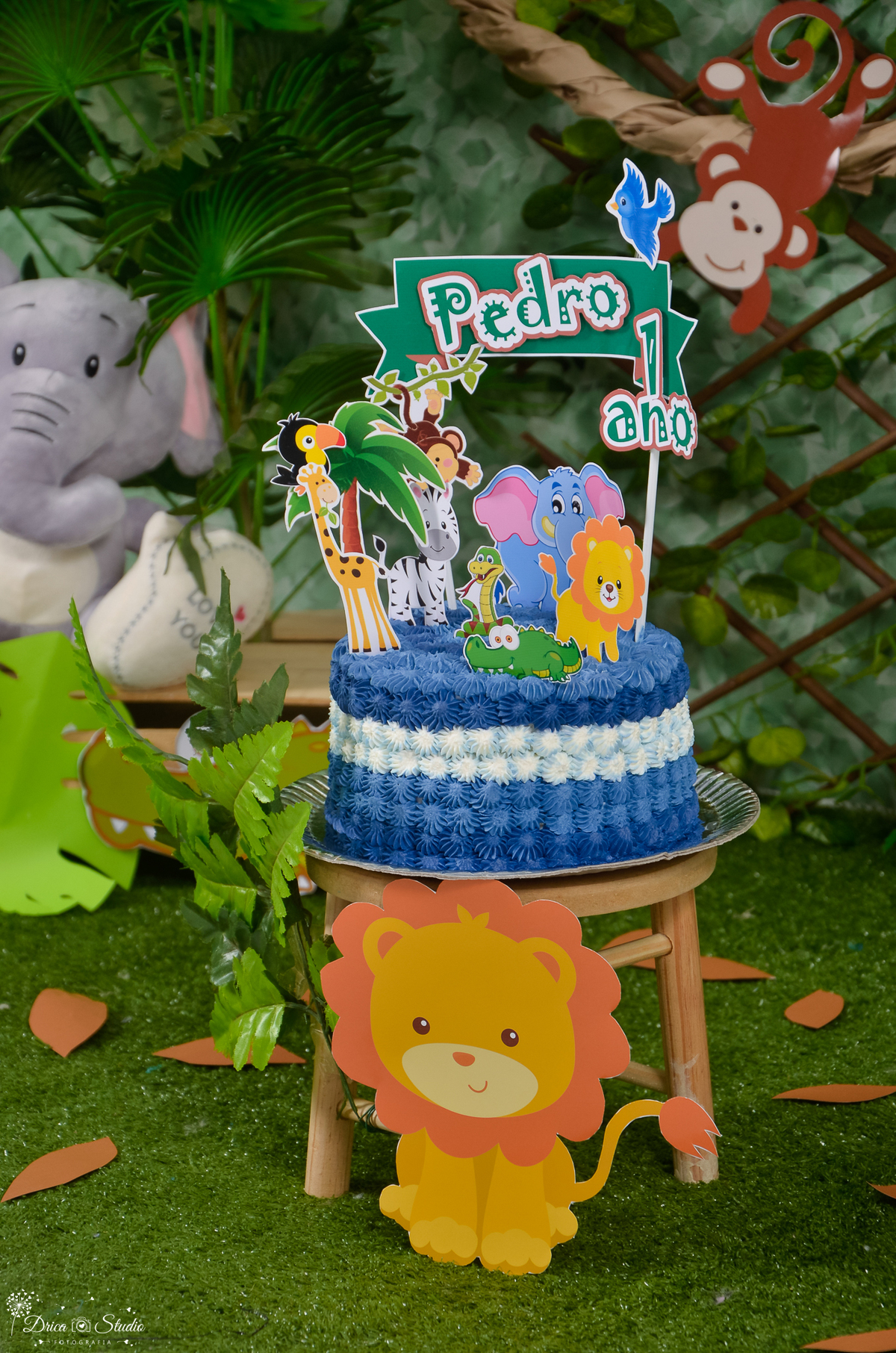 cenário Smash The Cake tema safári fundo verde com folhas, em cima de uma mini cadeira um bolo azul com plaquinhas de vários animais. Drica Studio Fotografias