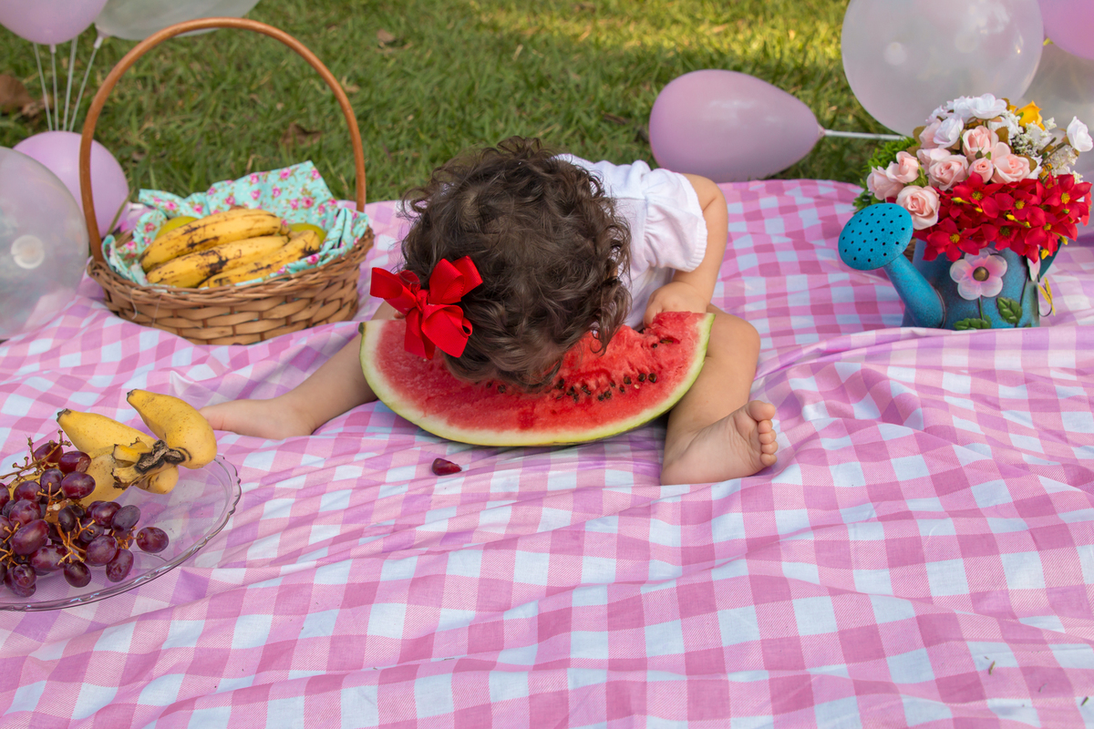 MENINA com LAÇO VERMELHO na cabeça sentada em uma toalha xadrez cor de rosa COMENDO de cara na MELANCIA no click da FOTÓGRAFA de ENSAIO SMASH THE FRUIT em SÃO PAULO, SP sorridente em uma imagem colorida