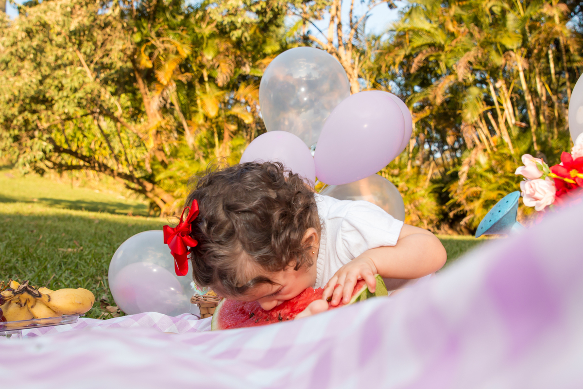 MENINA com LAÇO VERMELHO na cabeça sentada em uma toalha xadrez cor de rosa COMENDO de cara na MELANCIA no click da FOTÓGRAFA de ENSAIO SMASH THE FRUIT em SÃO PAULO, SP sorridente em uma imagem colorida