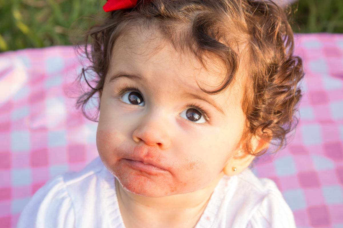 MENINA com LAÇO VERMELHO na cabeça sentada em uma toalha xadrez cor de rosa com a BOCA SUJA DE MELANCIA no click da FOTÓGRAFA de ENSAIO SMASH THE FRUIT em SÃO PAULO, SP sorridente em uma imagem colorida