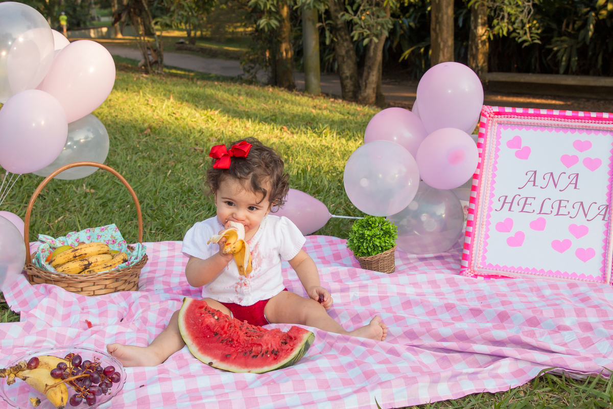 MENINA com LAÇO VERMELHO na cabeça sentada em uma toalha xadrez cor de rosa COMENDO BANANA no click da FOTÓGRAFA de ENSAIO SMASH THE FRUIT em SÃO PAULO, SP sorridente em uma imagem colorida