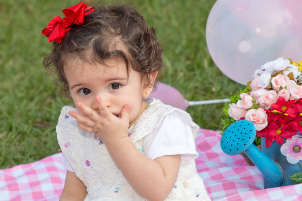 MENINA com LAÇO VERMELHO na cabeça sentada em uma toalha xadrez cor de rosa COMENDO e olhando para a CÂMERA no click da FOTÓGRAFA de ENSAIO SMASH THE FRUIT em SÃO PAULO, SP sorridente em uma imagem colorida