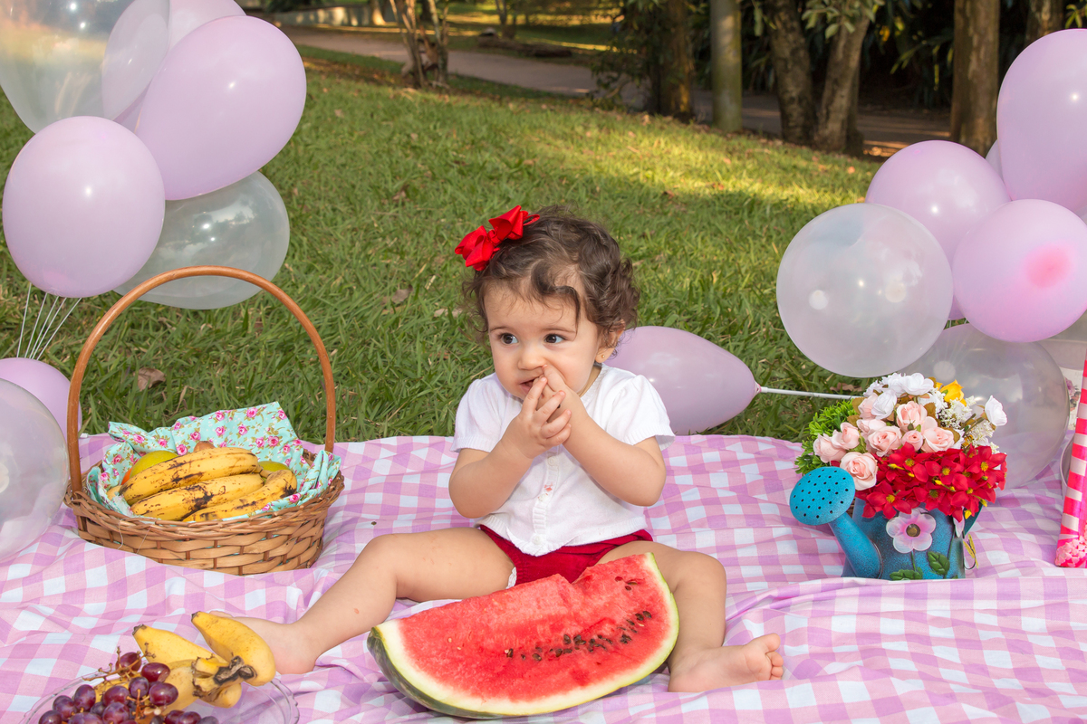 MENINA com LAÇO VERMELHO na cabeça sentada em uma toalha xadrez cor de rosa comendo MELANCIA no click da FOTÓGRAFA de ENSAIO SMASH THE FRUIT em SÃO PAULO, SP sorridente em uma imagem colorida