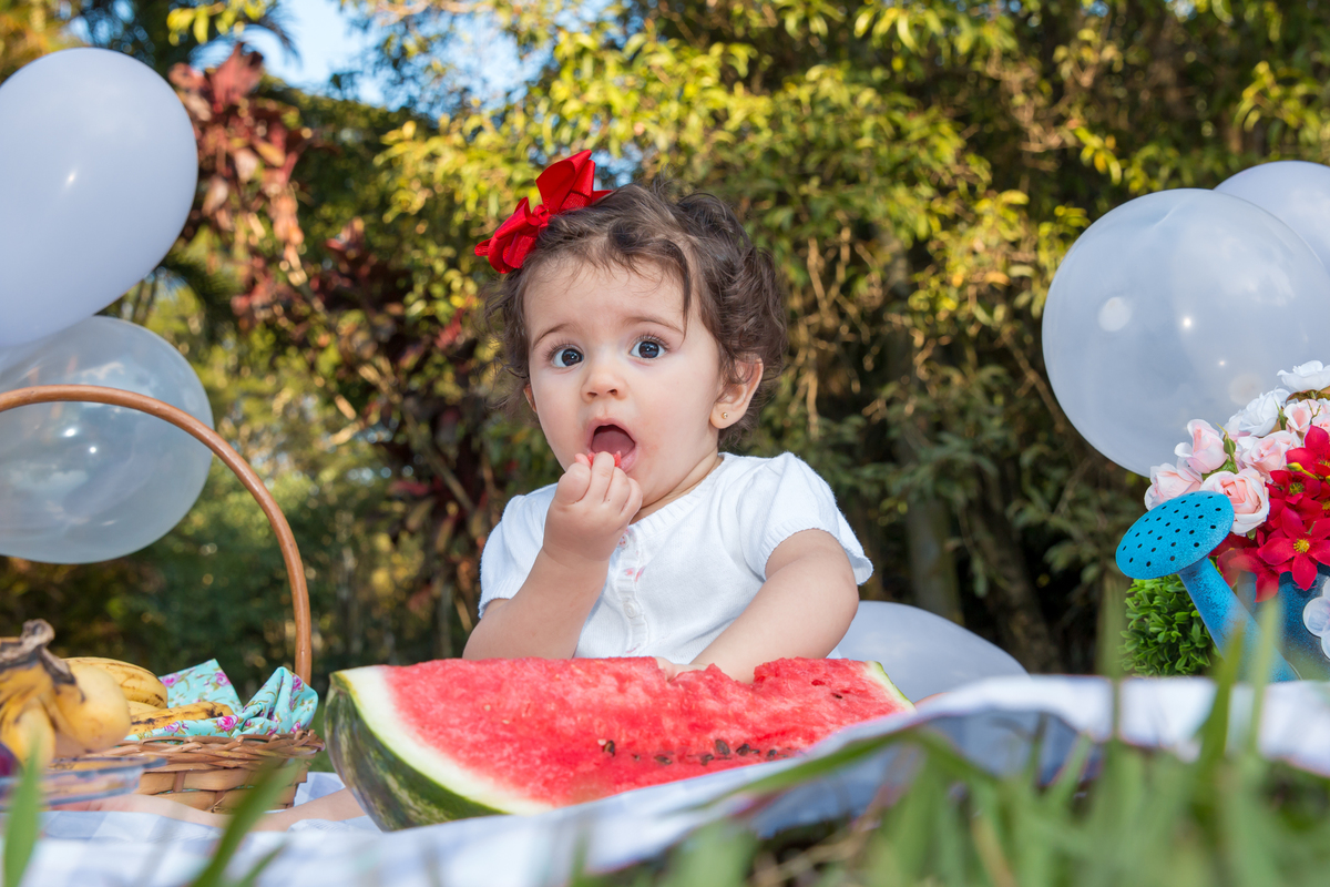 MENINA com LAÇO VERMELHO na cabeça sentada em uma toalha xadrez cor de rosa COMENDO MELANCIA e olhando para a CÂMERA no click da FOTÓGRAFA de ENSAIO SMASH THE FRUIT em SÃO PAULO, SP sorridente em uma imagem colorida