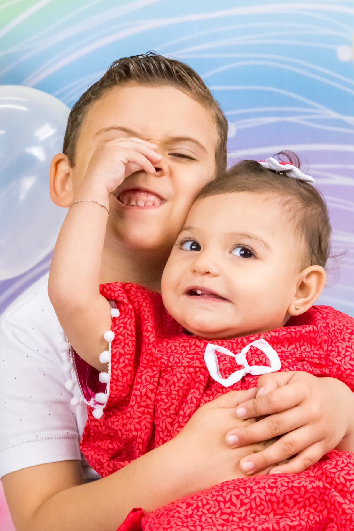 MENINA sentada de VESTIDO VERMELHO no colo do IRMÃO apertando o NARIZ do IRMÃO no click da FOTÓGRAFA de ENSAIO SMASH THE CAKE em SÃO PAULO, SP sorridentes em uma imagem colorida
