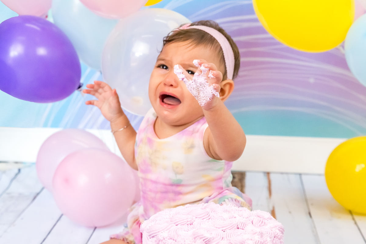 MENINA sentada CHORANDO com as MÃOS SUJAS DE BOLO para a CÂMERA no click da FOTÓGRAFA de ENSAIO SMASH THE CAKE em SÃO PAULO, SP sorridente em uma imagem colorida