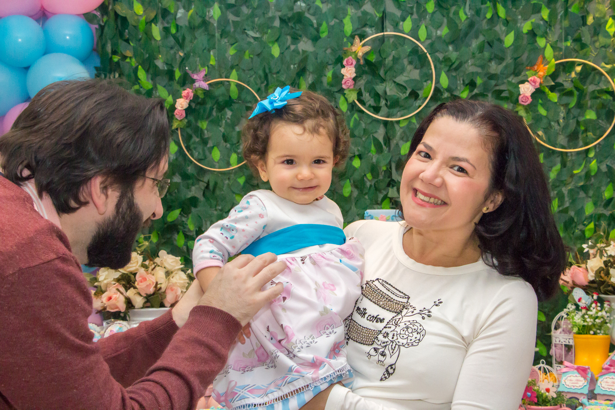 MENINA no colo dos PAIS de vestido com LAÇO AZUL em festa tema JARDIM ENCANTADO nas cores VERDE e CORES PASTÉIS no click da FOTÓGRAFA de FESTA INFANTIL em SÃO PAULO, SP em uma imagem colorida