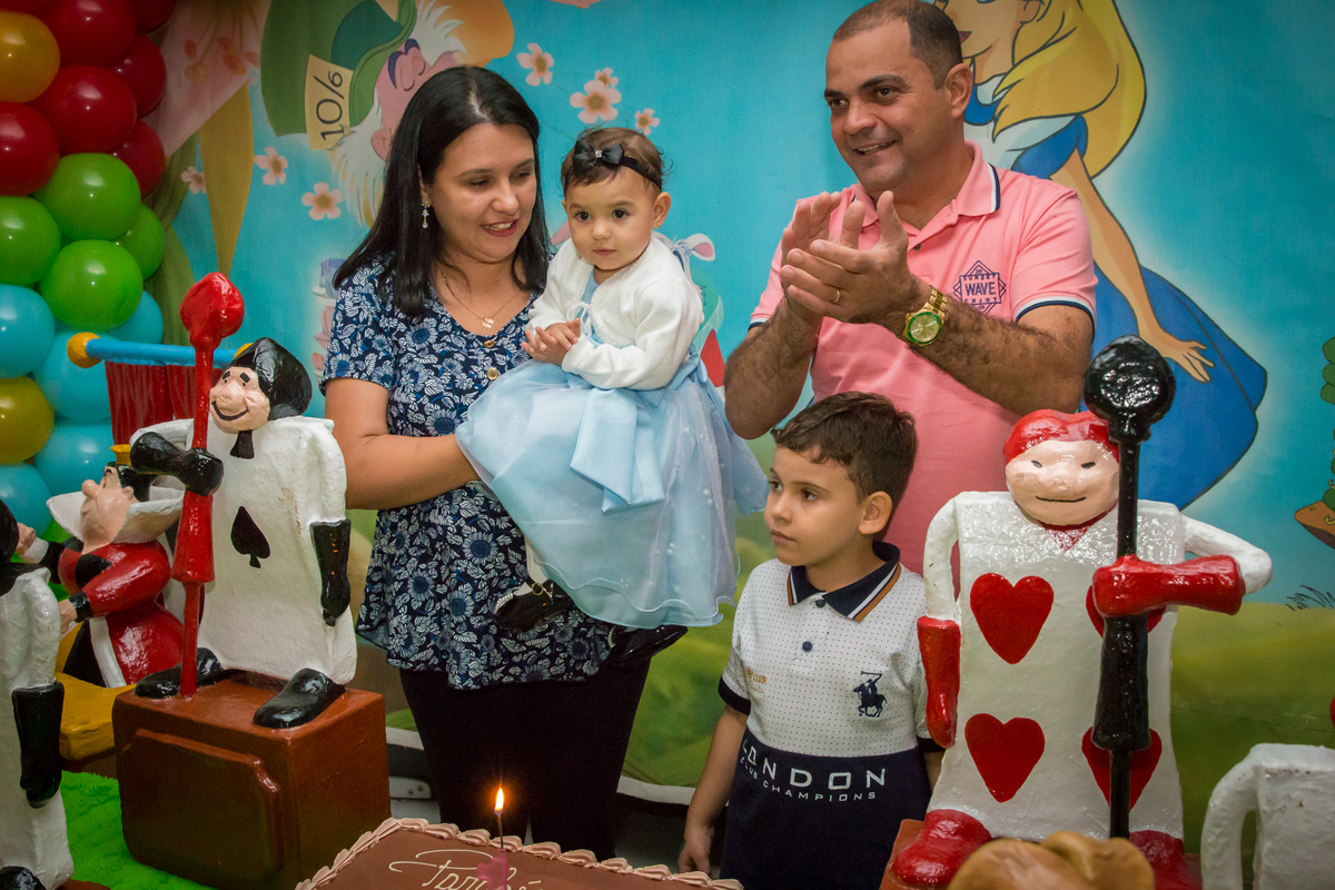 MENINA de VESTIDO AZUL no COLO dos PAIS cantando PARABÉNS em frente a decoração tema ALICE NO PÁIS DAS MARAVILHAS no click do FOTÓGRAFO de FESTA INFANTIL em SÃO PAULO, SP em uma imagem colorida