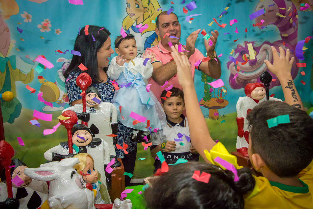 MENINA de VESTIDO AZUL no COLO dos PAIS cantando PARABÉNS em frente a decoração tema ALICE NO PÁIS DAS MARAVILHAS no click do FOTÓGRAFO de FESTA INFANTIL em SÃO PAULO, SP em uma imagem colorida