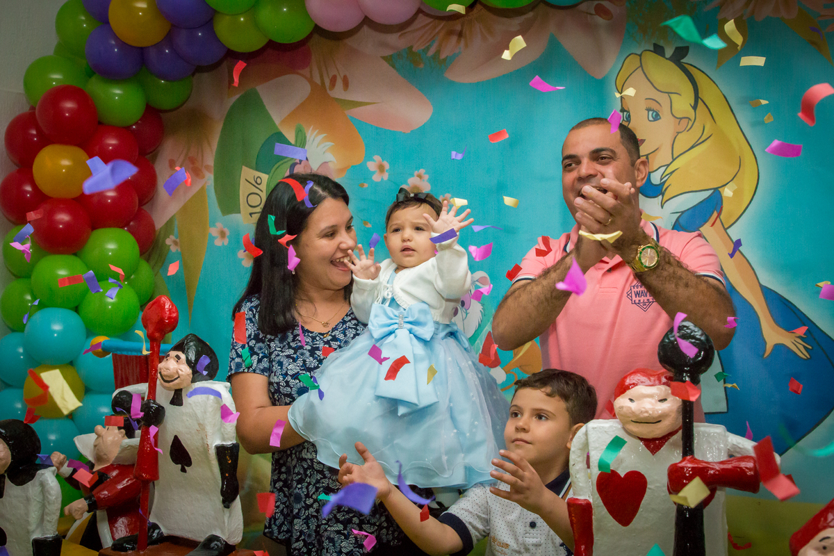 MENINA de VESTIDO AZUL no COLO dos PAIS cantando PARABÉNS em frente a decoração tema ALICE NO PÁIS DAS MARAVILHAS no click do FOTÓGRAFO de FESTA INFANTIL em SÃO PAULO, SP em uma imagem colorida