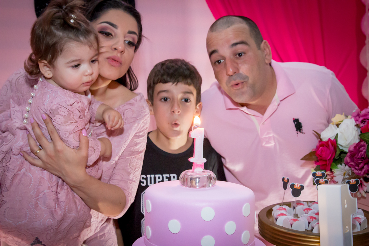 MENINA com a FAMÍLIA ASSOPRANDO a VELA em frente a decoração tema MINNIE nas cores ROSA e BRANCO no click do FOTÓGRAFO de FESTA INFANTIL em SÃO PAULO, SP em uma imagem colorida