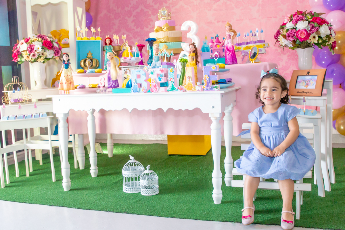 MENINA sorrindo de VESTIDO AZUL sentada em frente a decoração tema PRINCESAS nas cores ROSA, DOURADO e LILÁS no click do FOTÓGRAFO de FESTA INFANTIL em SÃO PAULO, SP em uma imagem colorida