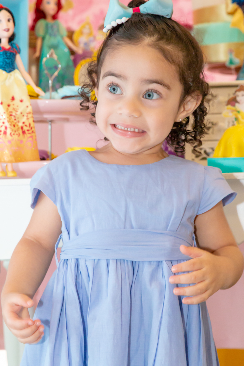 MENINA sorrindo de VESTIDO AZUL sentada em frente a decoração tema PRINCESAS nas cores ROSA, DOURADO e LILÁS no click do FOTÓGRAFO de FESTA INFANTIL em SÃO PAULO, SP em uma imagem colorida