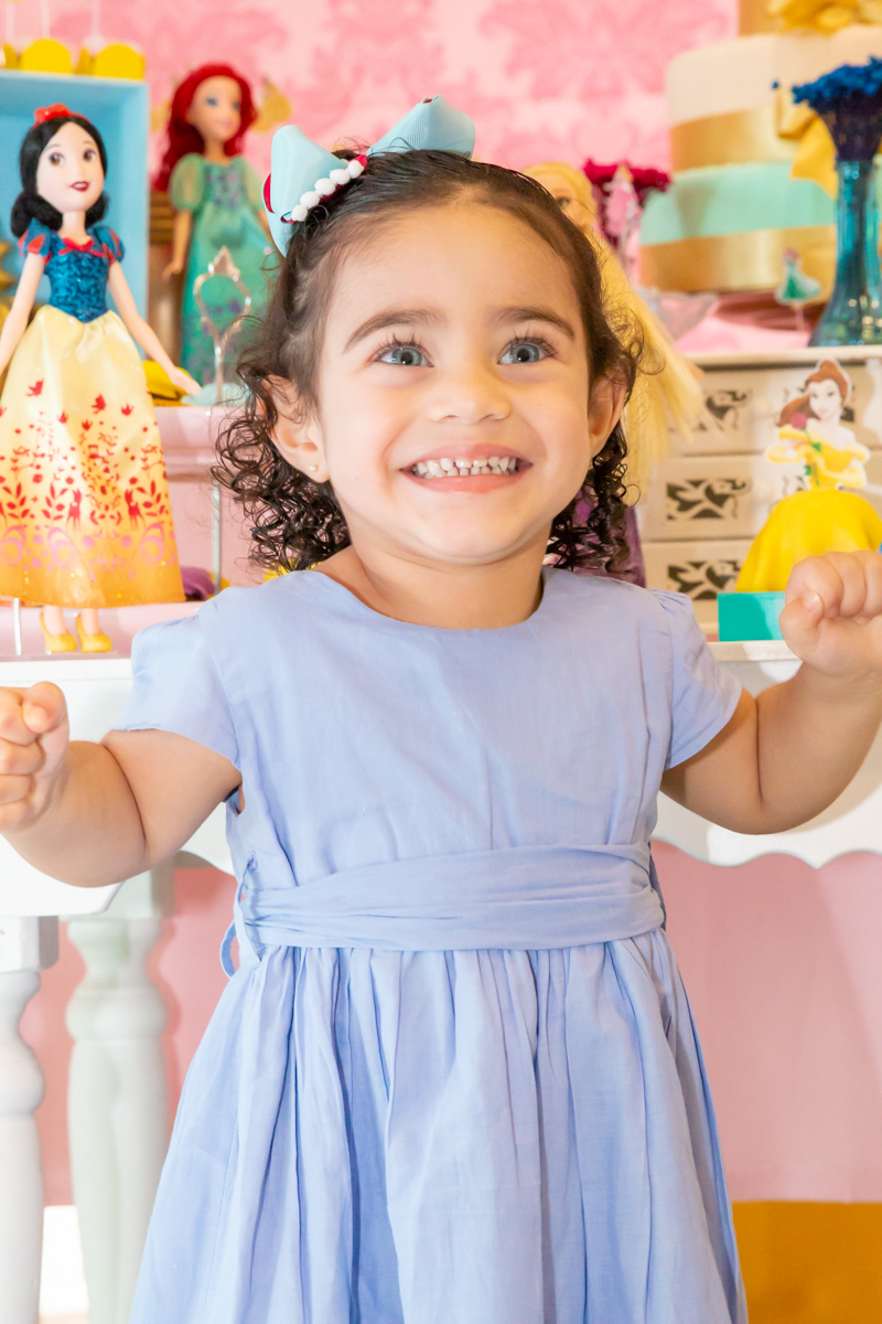 MENINA sorrindo de VESTIDO AZUL sentada em frente a decoração tema PRINCESAS nas cores ROSA, DOURADO e LILÁS no click do FOTÓGRAFO de FESTA INFANTIL em SÃO PAULO, SP em uma imagem colorida