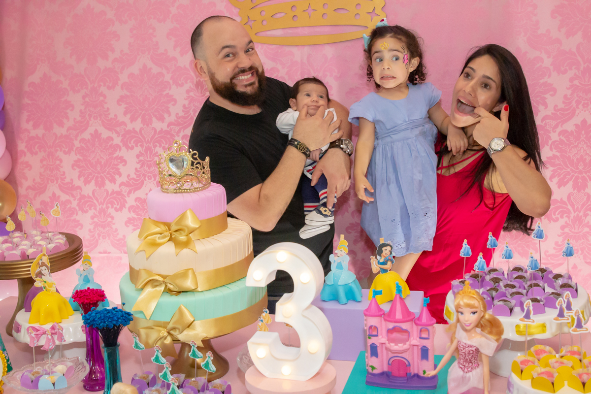 MENINA sorrindo de VESTIDO AZUL abraçada com os PAIS e IRMÃO em frente a decoração tema PRINCESAS nas cores ROSA, DOURADO e LILÁS no click do FOTÓGRAFO de FESTA INFANTIL em SÃO PAULO, SP em uma imagem colorida