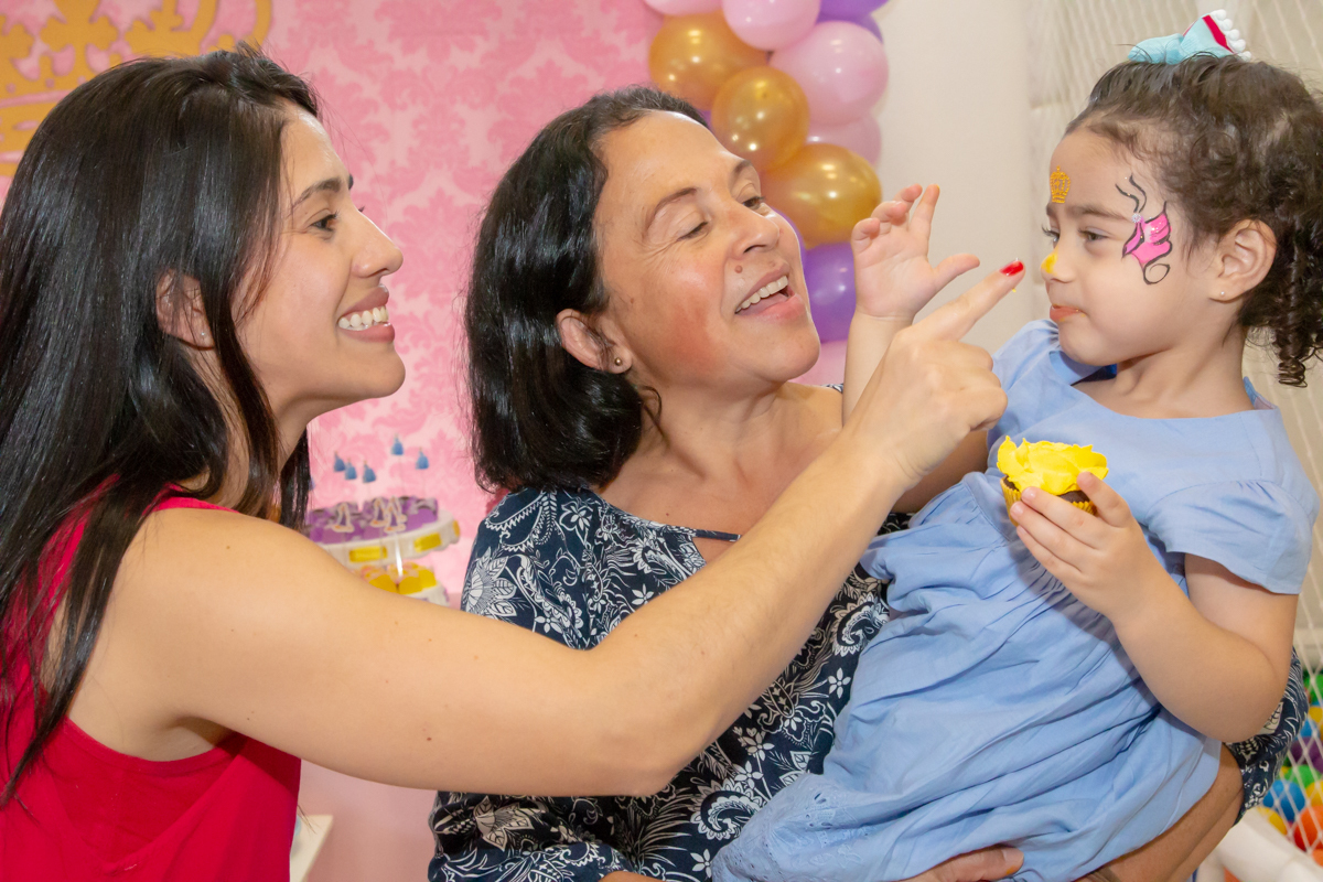 MENINA sorrindo de VESTIDO AZUL abraçada com MÃE e AVÓ em frente a decoração tema PRINCESAS nas cores ROSA, DOURADO e LILÁS no click do FOTÓGRAFO de FESTA INFANTIL em SÃO PAULO, SP em uma imagem colorida