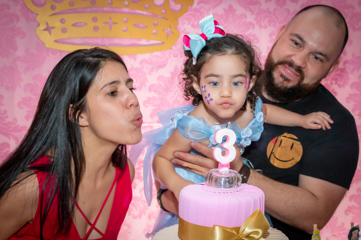 MENINA assoprando A VELA cantando PARABÉNS com os PAIS sorrindo de VESTIDO AZUL em decoração tema PRINCESAS nas cores ROSA, DOURADO e LILÁS no click do FOTÓGRAFO de FESTA INFANTIL em SÃO PAULO, SP em uma imagem colorida
