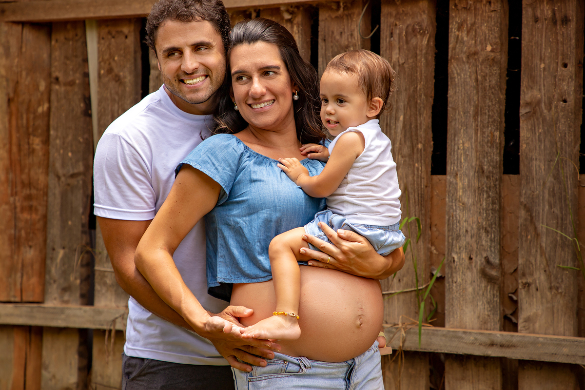 Mulher GRÁVIDA com a BARRIGA de fora, posando com o MARIDO e a FILHA em ENSAIO GESTANTE em um SÍTIO, em click do FOTÓGRAFO de GESTANTE.